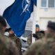 Soldier in camouflage holds a blue flag with a white emblem during a military ceremony, with colleagues in uniform nearby.