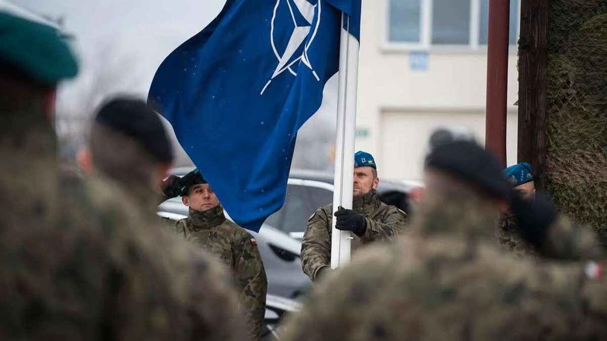 Soldier in camouflage holds a blue flag with a white emblem during a military ceremony, with colleagues in uniform nearby.