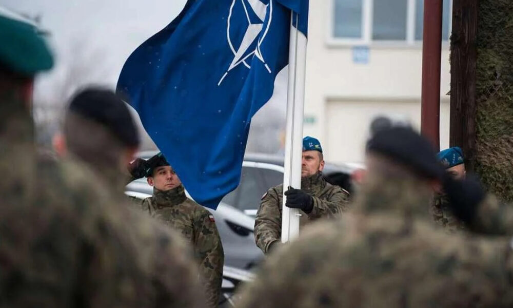 Soldier in camouflage holds a blue flag with a white emblem during a military ceremony, others stand in the foreground and background with blue berets.