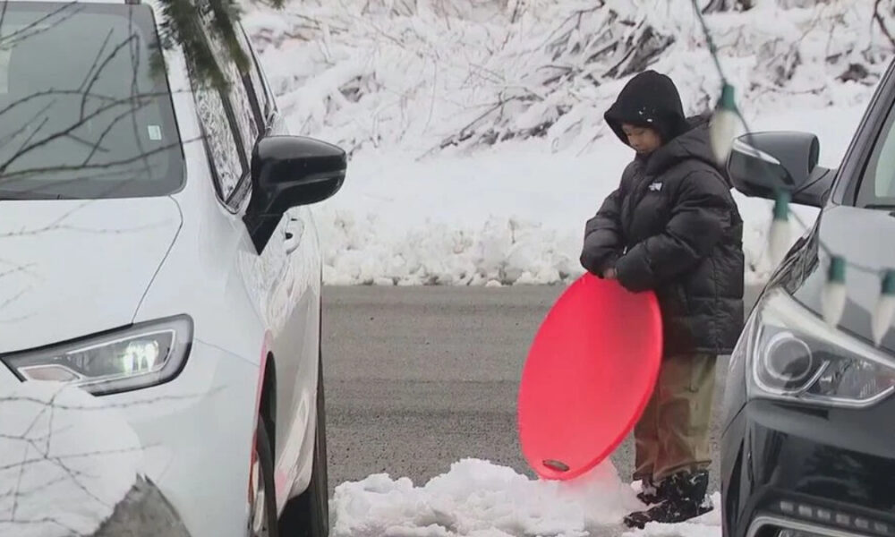 Child in a black hooded jacket standing between two cars in a snowy parking lot, holding a bright red round sled.