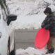 Child in a black hooded jacket standing between two cars in a snowy parking lot, holding a bright red round sled.