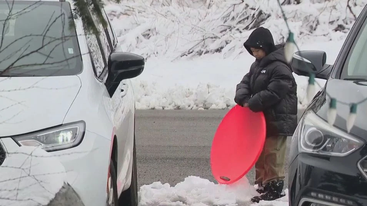 Child in a black hooded jacket standing between two cars in a snowy parking lot, holding a bright red round sled.