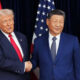 Two men in suits shake hands in front of US and China flags behind them.