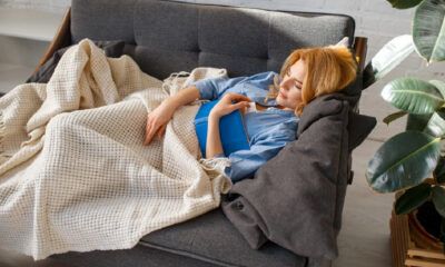 Woman lounging on a gray sofa, tucked in with a beige knit blanket and holding a blue book in a bright living room.