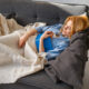 Woman lounging on a gray sofa, tucked in with a beige knit blanket and holding a blue book in a bright living room.