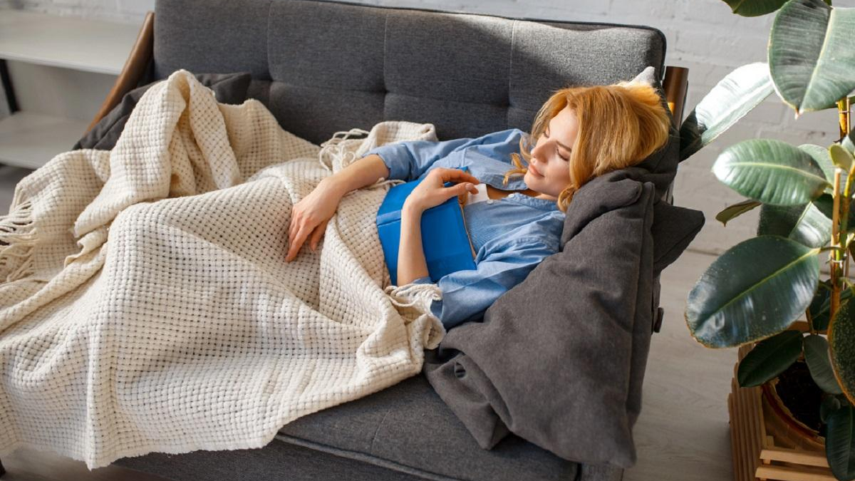 Woman lounging on a gray sofa, tucked in with a beige knit blanket and holding a blue book in a bright living room.