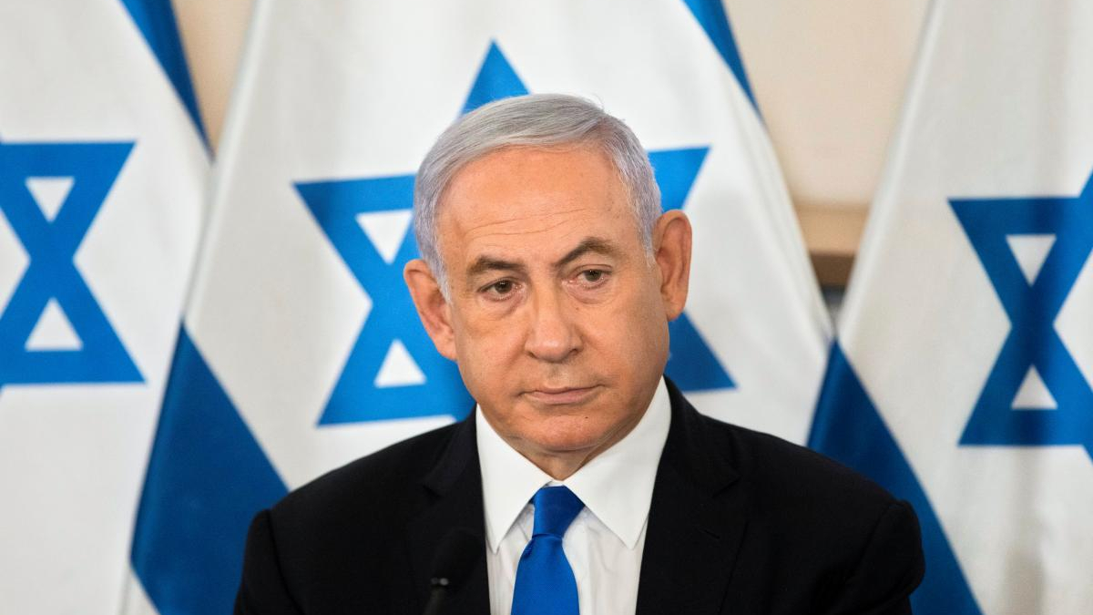 Man in a dark suit and blue tie speaks at a press event with Israeli flags in the background.
