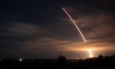 Night scene of a rocket launch with a bright diagonal exhaust trail across the cloudy sky and a glowing plume near the horizon, silhouetted trees below.