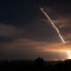 Night scene of a rocket launch with a bright diagonal exhaust trail across the cloudy sky and a glowing plume near the horizon, silhouetted trees below.