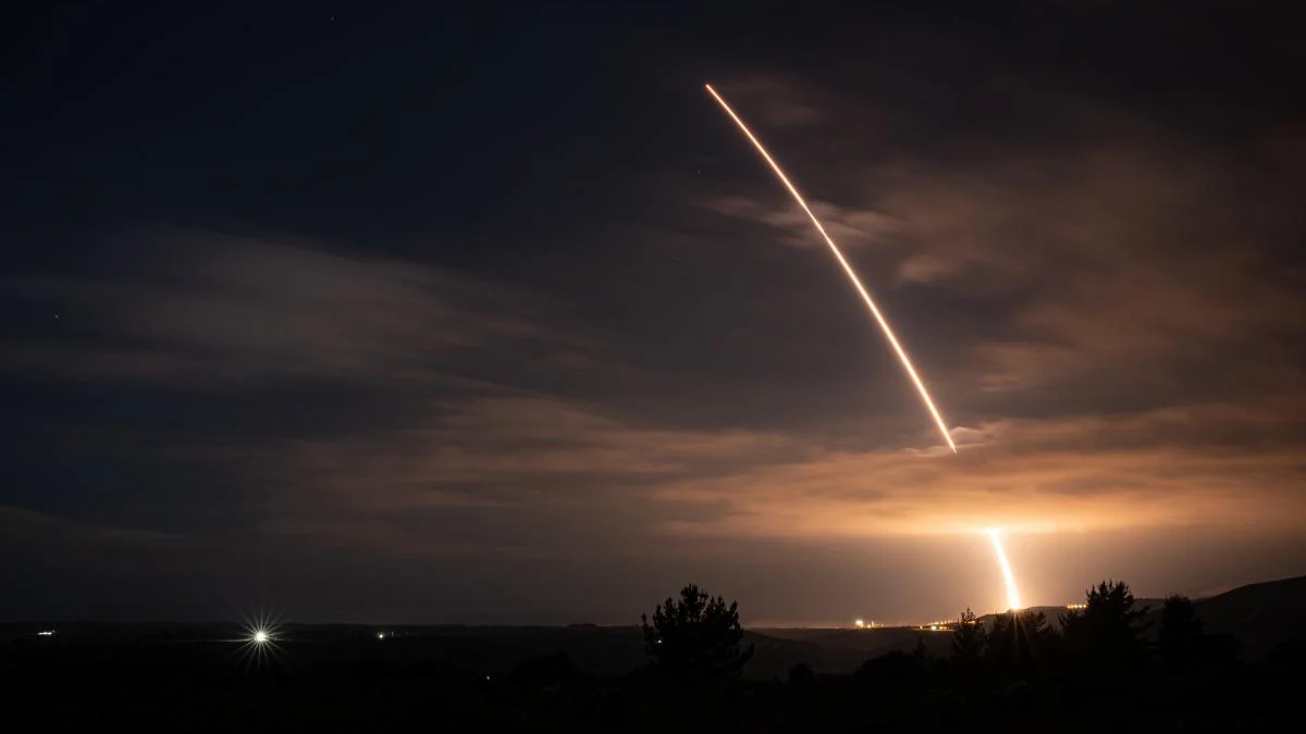 Night scene of a rocket launch with a bright diagonal exhaust trail across the cloudy sky and a glowing plume near the horizon, silhouetted trees below.
