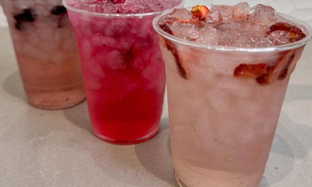 Three clear plastic cups with iced fruit drinks on a light countertop; front cup is pale pink with fruit slices and crushed ice, others are red and brownish.
