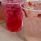 Three clear plastic cups with iced fruit drinks on a light countertop; front cup is pale pink with fruit slices and crushed ice, others are red and brownish.