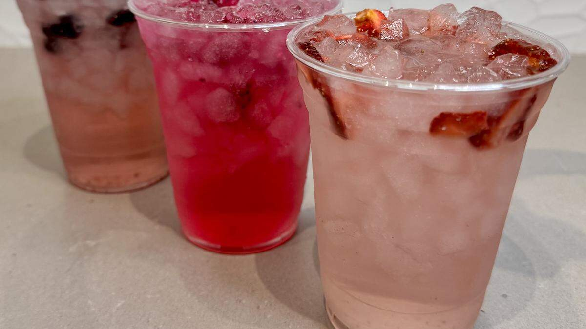 Three clear plastic cups with iced fruit drinks on a light countertop; front cup is pale pink with fruit slices and crushed ice, others are red and brownish.