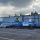 Boot Barn storefront under construction, construction equipment behind a chain-link fence in a parking lot under a cloudy sky.