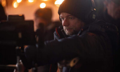 Cameraman wearing a black beanie and headphones adjusting a camera in a dim, warm-lit set with blurred background lights behind him.