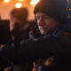 Cameraman wearing a black beanie and headphones adjusting a camera in a dim, warm-lit set with blurred background lights behind him.
