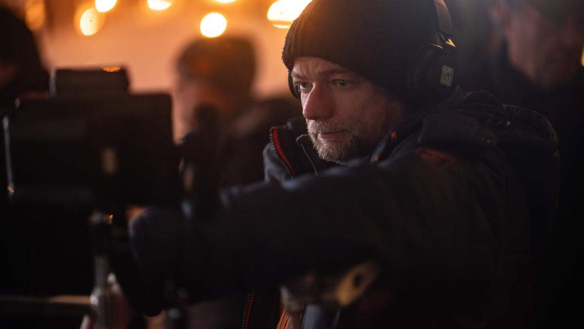 Cameraman wearing a black beanie and headphones adjusting a camera in a dim, warm-lit set with blurred background lights behind him.
