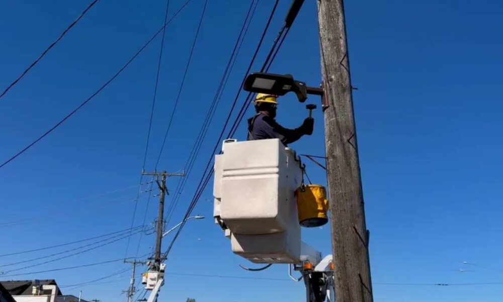 Utility worker in a bucket lift repairing a streetlight on a wooden utility pole against a clear blue sky, with power lines in the background.