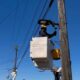 Utility worker in a bucket lift repairing a streetlight on a wooden utility pole against a clear blue sky, with power lines in the background.