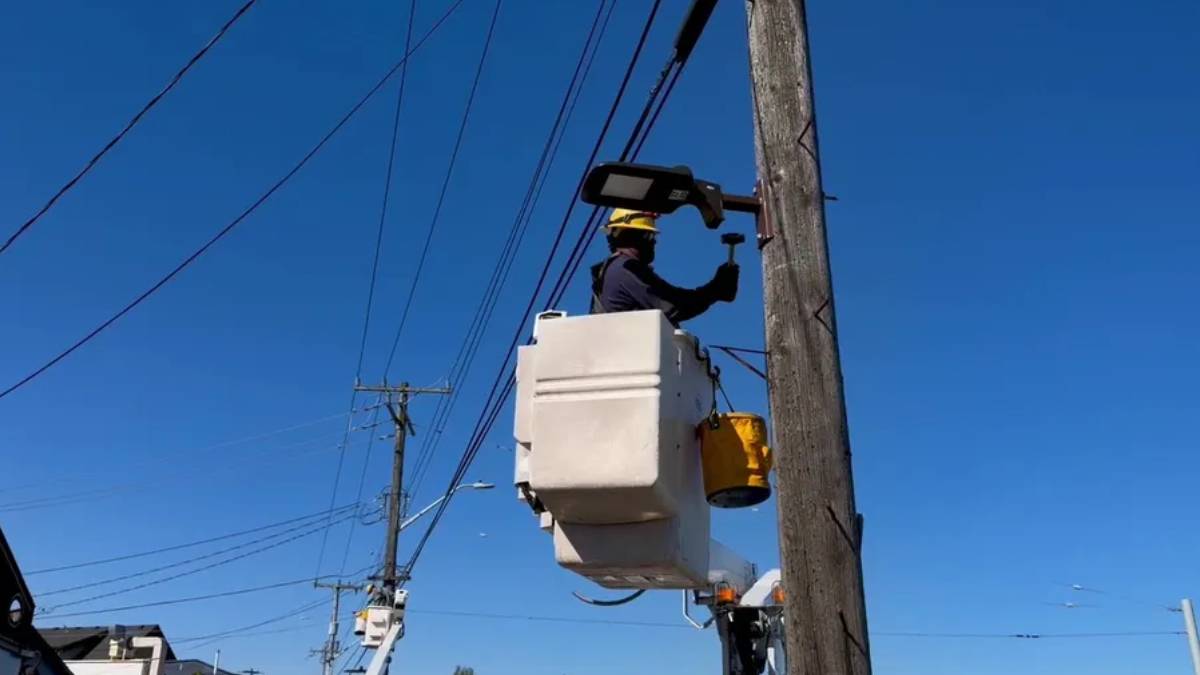 Utility worker in a bucket lift repairing a streetlight on a wooden utility pole against a clear blue sky, with power lines in the background.