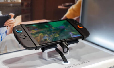 Nintendo Switch 2 handheld game console on a display stand, screen showing a colorful underwater-style game, with people blurred in the background.