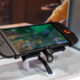Nintendo Switch 2 handheld game console on a display stand, screen showing a colorful underwater-style game, with people blurred in the background.