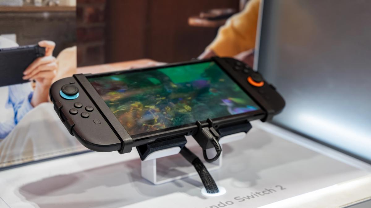 Nintendo Switch 2 handheld game console on a display stand, screen showing a colorful underwater-style game, with people blurred in the background.