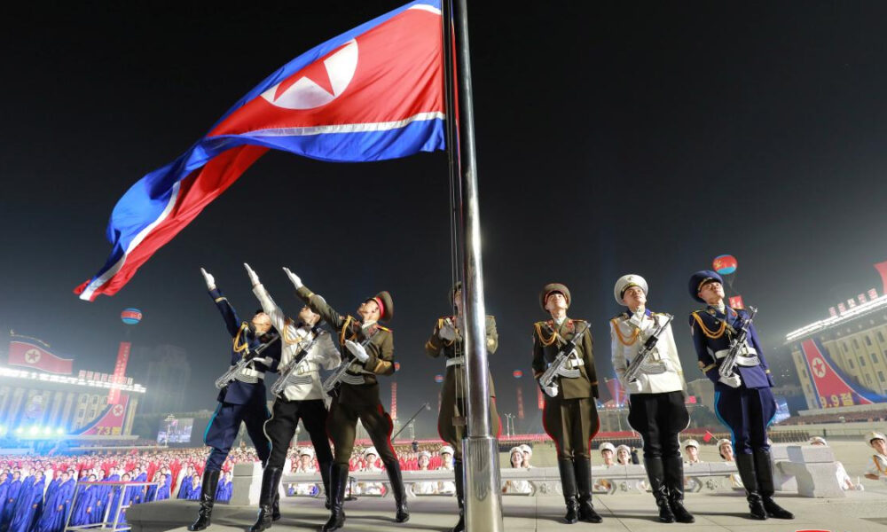 Nighttime military flag-raising ceremony with uniformed soldiers saluting as a large red flag is raised on a tall pole.
