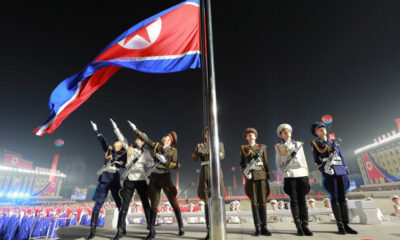 Nighttime military flag-raising ceremony with uniformed soldiers saluting as a large red flag is raised on a tall pole.