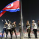 Nighttime military flag-raising ceremony with uniformed soldiers saluting as a large red flag is raised on a tall pole.