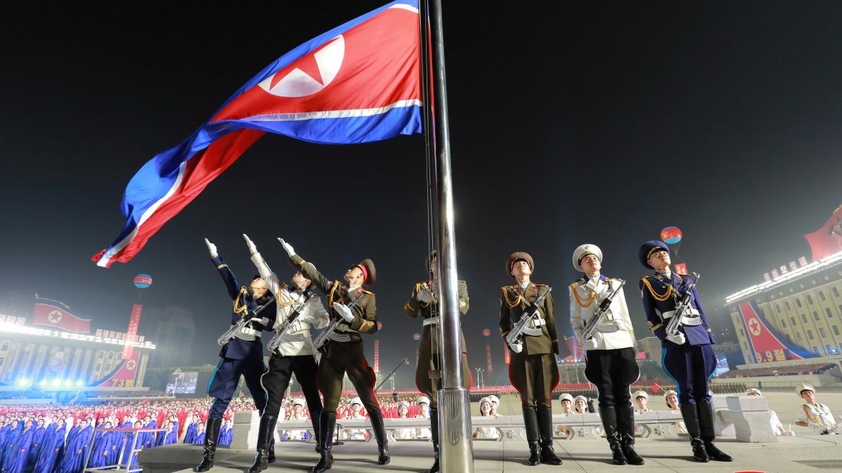 Nighttime military flag-raising ceremony with uniformed soldiers saluting as a large red flag is raised on a tall pole.