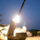 Mobile missile launcher on tracks fires a rocket into the dark sky, with bright exhaust and smoke surrounding the platform.