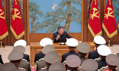 Kim Jong Un speaks at a wooden podium in a formal hall with red flags beside him and a blue mural behind