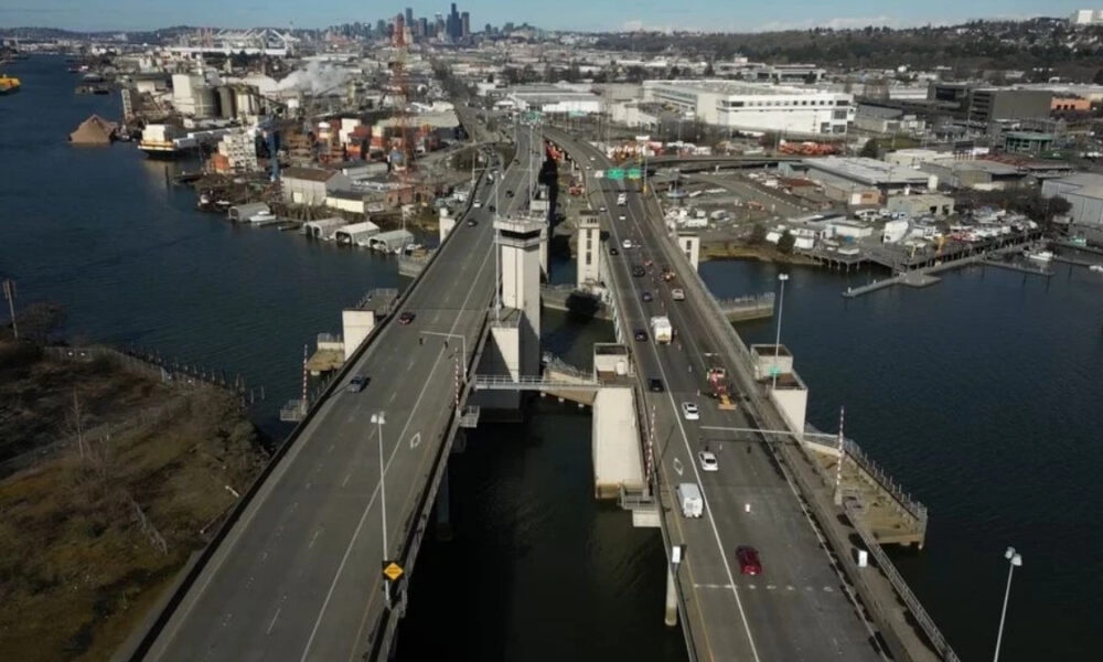 Aerial view of a large bridge over a wide river with industrial buildings and a distant city skyline on the horizon.