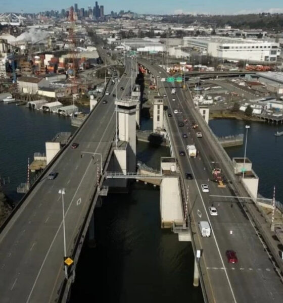 Aerial view of a large bridge over a wide river with industrial buildings and a distant city skyline on the horizon.