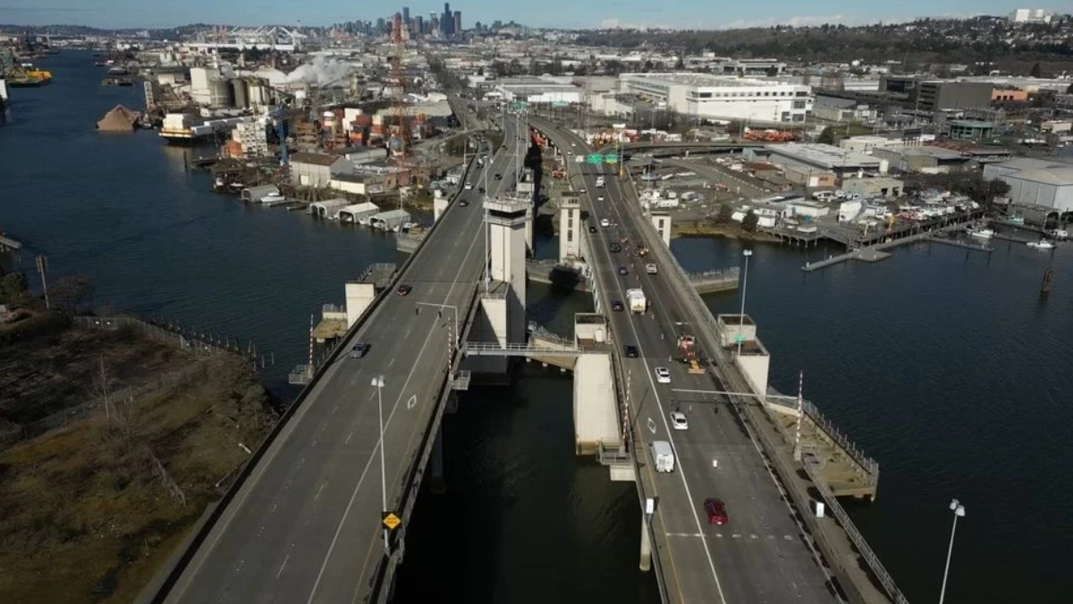 Aerial view of a large bridge over a wide river with industrial buildings and a distant city skyline on the horizon.