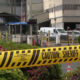 Yellow 'Do Not Cross' and 'Caution: Sheriff Line' tape in the foreground with a gated area and vehicles in the background, suggesting a restricted scene.