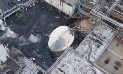 Aerial view of an industrial plant with a toppled circular tank, dark liquid on the ground and scattered debris among pipes and walkways.