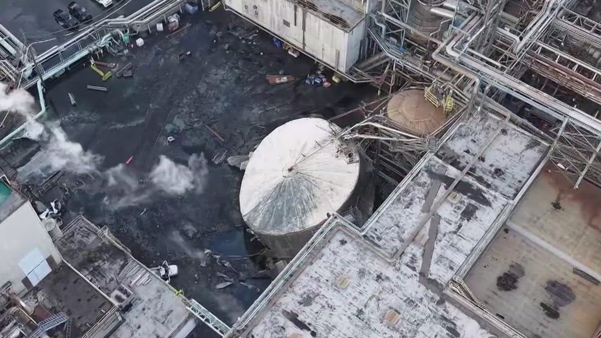Aerial view of an industrial plant with a toppled circular tank, dark liquid on the ground and scattered debris among pipes and walkways.