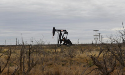 Oil pump jack operating in a dry, desert-like field with scrubby brush and distant power lines.