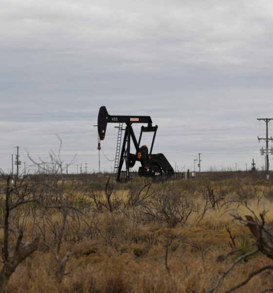 Oil pump jack operating in a dry, desert-like field with scrubby brush and distant power lines.