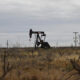 Oil pump jack operating in a dry, desert-like field with scrubby brush and distant power lines.