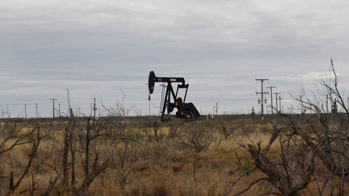 Oil pump jack operating in a dry, desert-like field with scrubby brush and distant power lines.