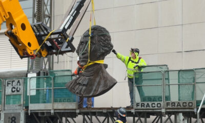 Bronze bust sculpture being lifted by a crane as workers guide it on a construction platform.