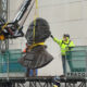 Bronze bust sculpture being lifted by a crane as workers guide it on a construction platform.