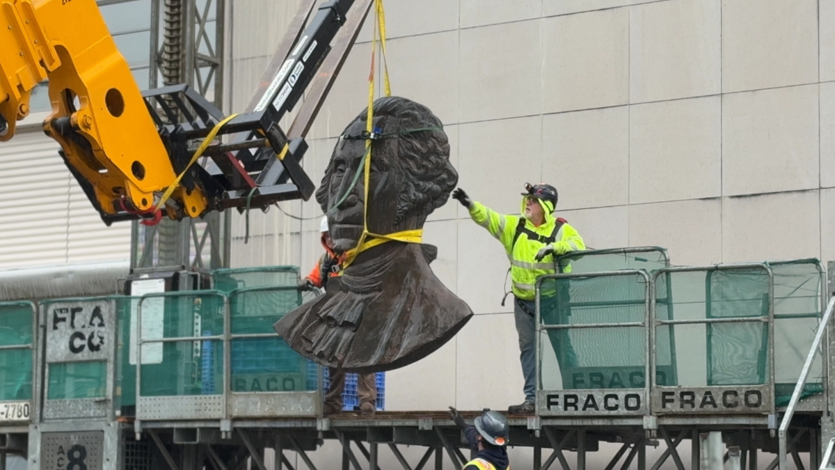 Bronze bust sculpture being lifted by a crane as workers guide it on a construction platform.