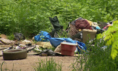 Pile of discarded trash and furniture—tires, bags, clothing, and a chair—dumped in a dense, overgrown outdoor area.