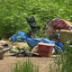 Pile of discarded trash and furniture—tires, bags, clothing, and a chair—dumped in a dense, overgrown outdoor area.