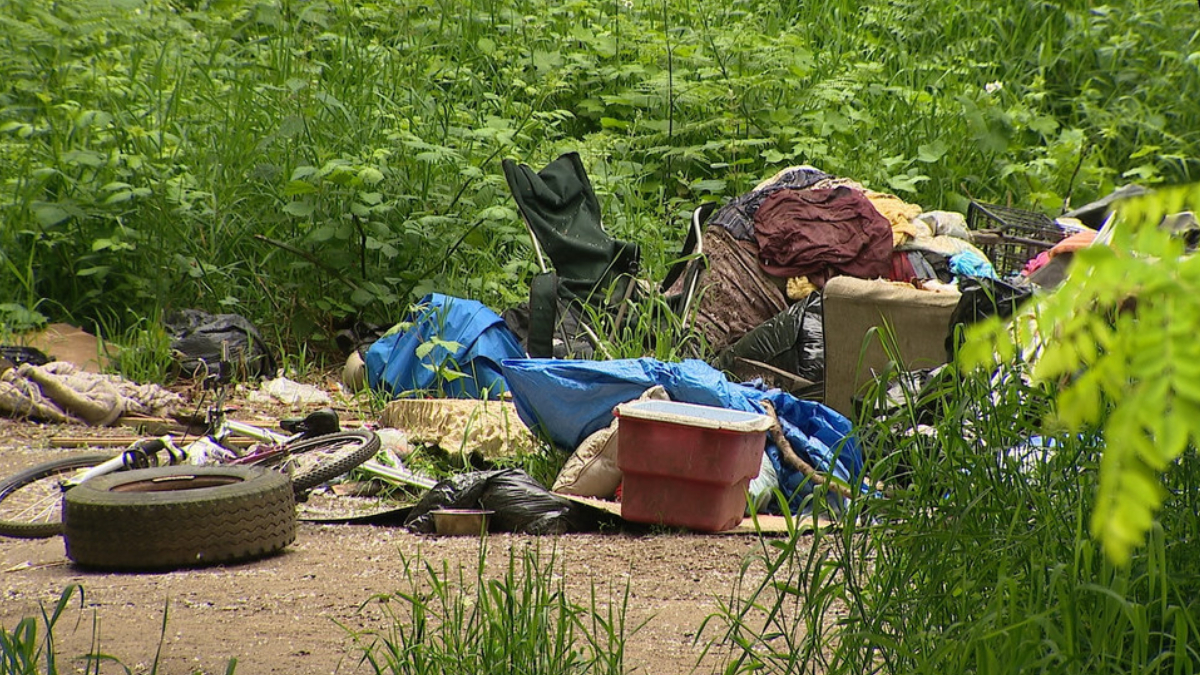 Pile of discarded trash and furniture—tires, bags, clothing, and a chair—dumped in a dense, overgrown outdoor area.
