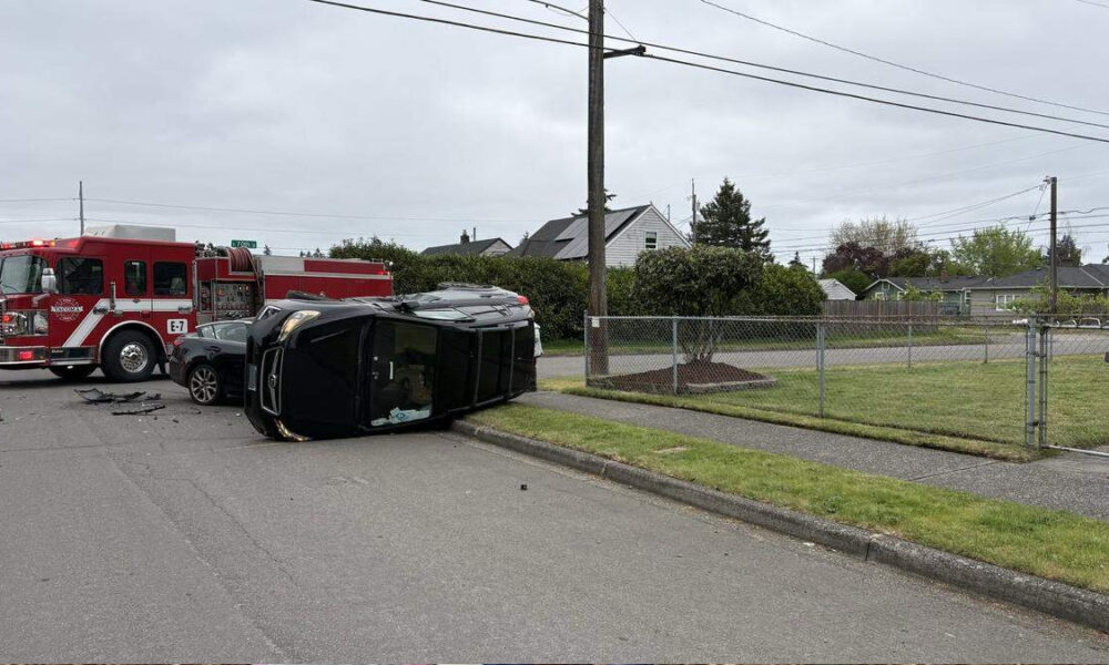 Black SUV resting on its side after a crash, with a red fire truck and firefighters at the scene in a residential street setting.